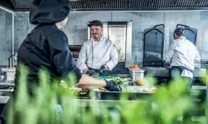 Un chef sonriente, un entremetier en cocina, prepara ingredientes mientras otro colega trabaja en el fondo.