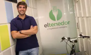 Hombre sonriente apoyado en pared colorida, con logo de El Tenedor, empresa de TripAdvisor, y una bicicleta.