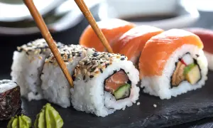Palillos recogen un rollo de sushi con salmón y pepino, mostrando cómo se hace el sushi. Otros rollos con salmón y wasabi al lado.