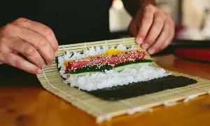Manos expertas demuestran como se enrolla el sushi, colocando atún, pepino y pimiento sobre arroz y alga nori en una esterilla de bambú.