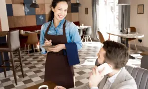 Camarera sonriente toma nota de un pedido, mostrando los diversos tipos de servicios en restaurantes. Un cliente habla por teléfono.