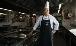 Un cocinero con gorro blanco y delantal negro posa en una cocina profesional.