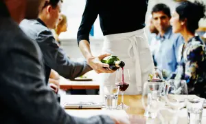 Camarero sirviendo vino tinto en una copa, mientras los comensales esperan para desbarasar una mesa y continuar la velada.
