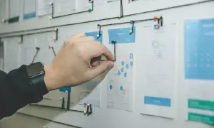 Un tablero visual con un mapa y wireframes de app, conectado con hilos. Una mano ajusta un hilo, demostrando la organización y flujo de trabajo que busca el **lean management**.