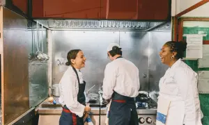 Tres chefs ríen en una cocina profesional, aprendiendo técnicas de cocina italiana.