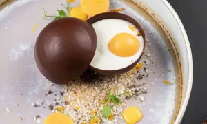 Delicioso trampantojo de comida: una esfera de chocolate oscuro abierta revela un postre cremoso con una yema simulada, rodeado de coco rallado y detalles naranjas.