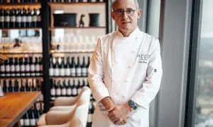 Chef Paco Pérez, con su chaquetilla blanca, posa con confianza en un restaurante elegante, rodeado de botellas de vino.