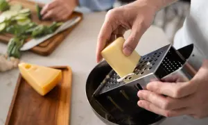 Manos rallando queso para una deliciosa comida. En el fondo, se preparan verduras.