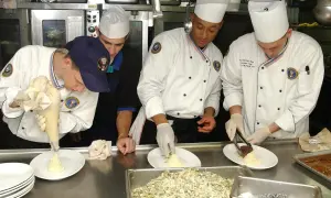 Un ayudante de cocina usa una manga pastelera para decorar un plato. Otros cocineros preparan la comida en una cocina profesional.
