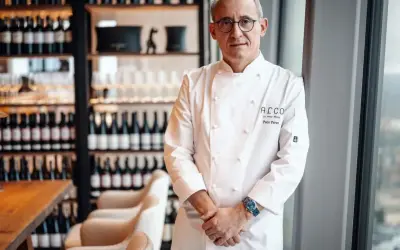 Chef Paco Pérez, con su chaquetilla blanca, posa con confianza en un restaurante elegante, rodeado de botellas de vino.