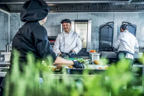 Un chef sonriente, un entremetier en cocina, prepara ingredientes mientras otro colega trabaja en el fondo.