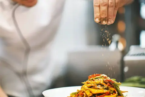 Un chef con título de cocinero espolvorea especias sobre un plato de verduras salteadas.