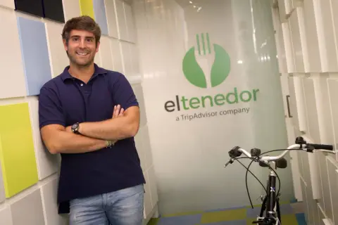 Hombre sonriente apoyado en pared colorida, con logo de El Tenedor, empresa de TripAdvisor, y una bicicleta.