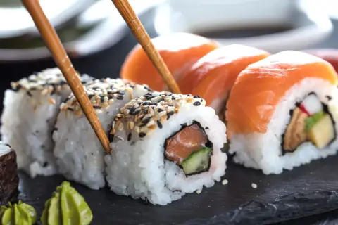Palillos recogen un rollo de sushi con salmón y pepino, mostrando cómo se hace el sushi. Otros rollos con salmón y wasabi al lado.