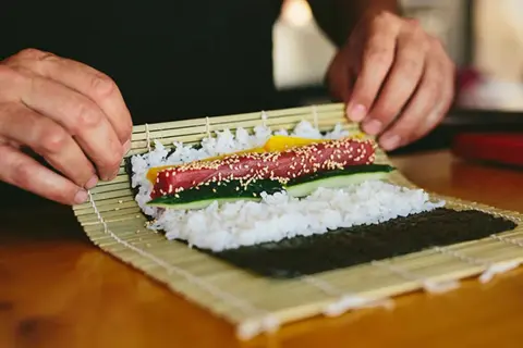 Manos expertas demuestran como se enrolla el sushi, colocando atún, pepino y pimiento sobre arroz y alga nori en una esterilla de bambú.