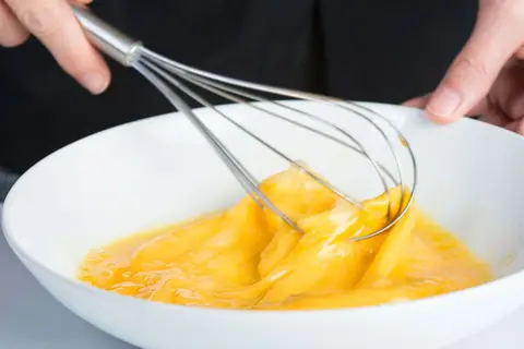 Manos batiendo huevos en un bol blanco, un paso clave para que es emulsionar y lograr texturas perfectas en la cocina.