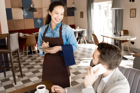 Camarera sonriente toma nota de un pedido, mostrando los diversos tipos de servicios en restaurantes. Un cliente habla por teléfono.