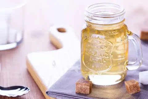 Jar de vidrio con líquido dorado, ideal para tu azucar invertida receta. Cubos de azúcar y tabla de madera al fondo.