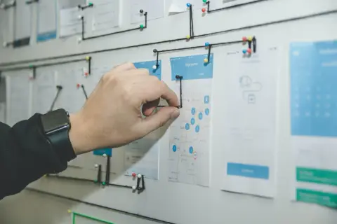 Un tablero visual con un mapa y wireframes de app, conectado con hilos. Una mano ajusta un hilo, demostrando la organización y flujo de trabajo que busca el **lean management**.