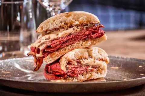 Mollete de pastrami con abundante carne curada y salsa cremosa, apilado en un plato oscuro.