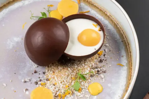 Delicioso trampantojo de comida: una esfera de chocolate oscuro abierta revela un postre cremoso con una yema simulada, rodeado de coco rallado y detalles naranjas.