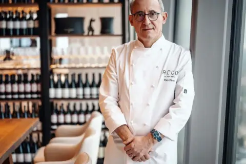 Chef Paco Pérez, con su chaquetilla blanca, posa con confianza en un restaurante elegante, rodeado de botellas de vino.