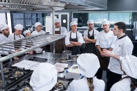 Estudiantes en un curso gestión restaurantes gratis, atentos a las instrucciones de un chef en una cocina profesional.