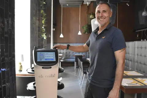Un hombre sonriente junto a un robot que sirve comida en un restaurante moderno.