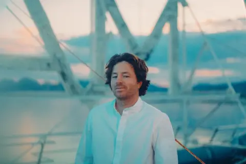 Jean Imbert, con camisa blanca, posa frente a una estructura de barco al atardecer.