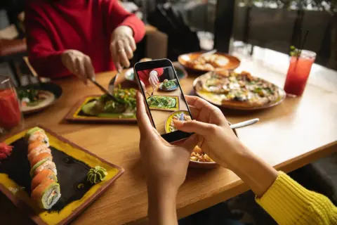 Persona toma foto de su comida en un restaurante para compartir en redes sociales. Se ven sushi, pizza y ensalada.