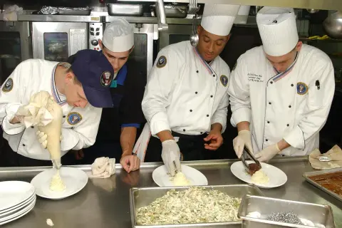 Un ayudante de cocina usa una manga pastelera para decorar un plato. Otros cocineros preparan la comida en una cocina profesional.