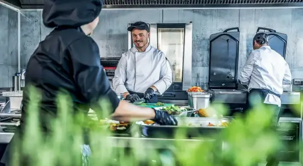 Un chef sonriente, un entremetier en cocina, prepara ingredientes mientras otro colega trabaja en el fondo.