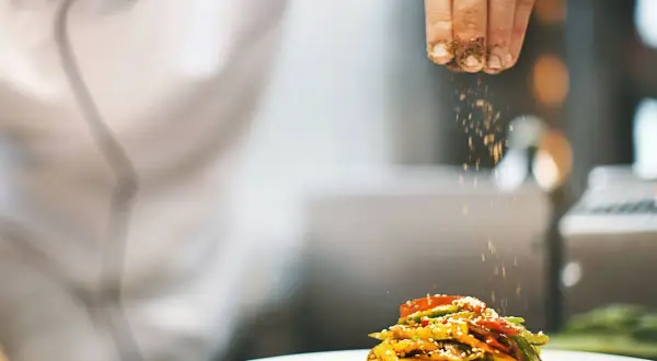 Un chef con título de cocinero espolvorea especias sobre un plato de verduras salteadas.