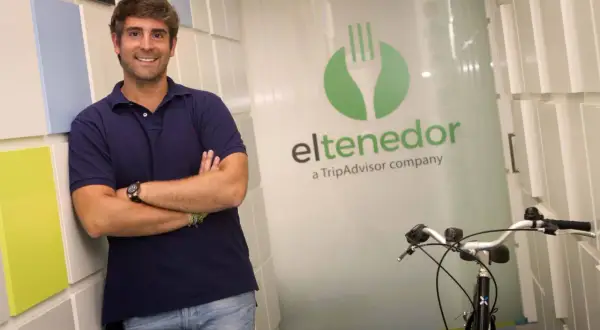 Hombre sonriente apoyado en pared colorida, con logo de El Tenedor, empresa de TripAdvisor, y una bicicleta.