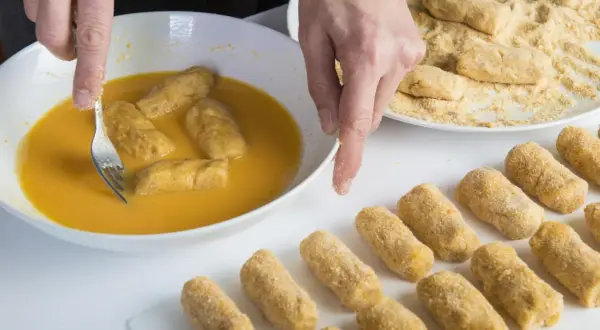 Manos rebozando croquetas en huevo. Se puede congelar la masa de croquetas para freír después.