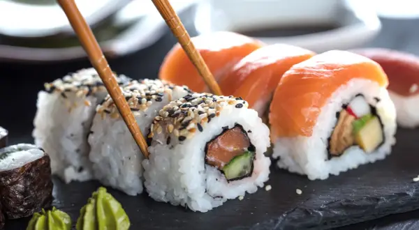 Palillos recogen un rollo de sushi con salmón y pepino, mostrando cómo se hace el sushi. Otros rollos con salmón y wasabi al lado.