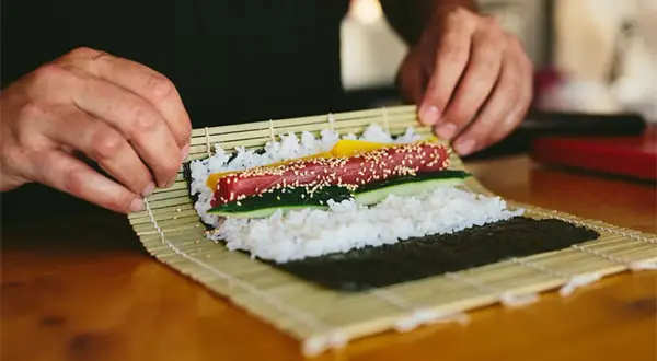 Manos expertas demuestran como se enrolla el sushi, colocando atún, pepino y pimiento sobre arroz y alga nori en una esterilla de bambú.