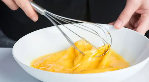 Manos batiendo huevos en un bol blanco, un paso clave para que es emulsionar y lograr texturas perfectas en la cocina.