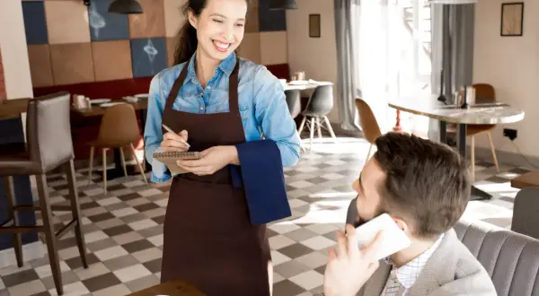 Camarera sonriente toma nota de un pedido, mostrando los diversos tipos de servicios en restaurantes. Un cliente habla por teléfono.