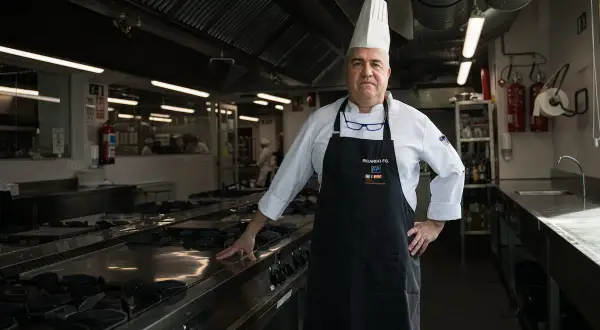 Un cocinero con gorro blanco y delantal negro posa en una cocina profesional.