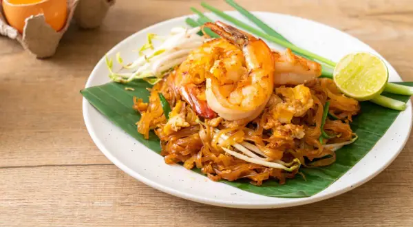 Pad Thai con camarones, perfecto para tu curso de cocina tailandesa. Servido sobre hoja de plátano, con brotes de soja, cebolleta y limón.