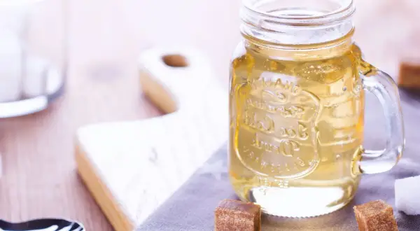 Jar de vidrio con líquido dorado, ideal para tu azucar invertida receta. Cubos de azúcar y tabla de madera al fondo.