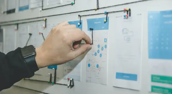 Un tablero visual con un mapa y wireframes de app, conectado con hilos. Una mano ajusta un hilo, demostrando la organización y flujo de trabajo que busca el **lean management**.