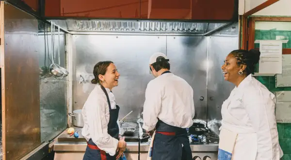 Tres chefs ríen en una cocina profesional, aprendiendo técnicas de cocina italiana.