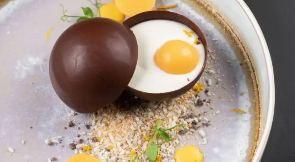 Delicioso trampantojo de comida: una esfera de chocolate oscuro abierta revela un postre cremoso con una yema simulada, rodeado de coco rallado y detalles naranjas.