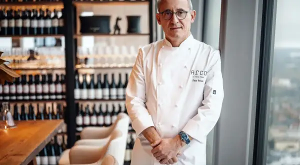 Chef Paco Pérez, con su chaquetilla blanca, posa con confianza en un restaurante elegante, rodeado de botellas de vino.