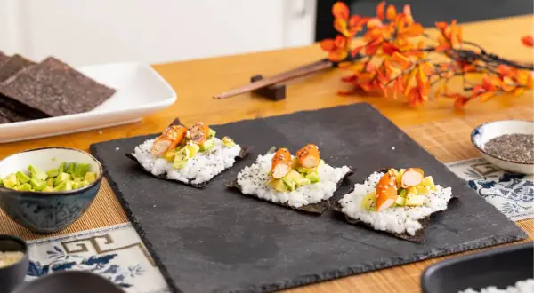 Tres pequeños rollos de sushi en proceso, listos para aprender a hacer sushi. Arroz, aguacate y palitos de cangrejo sobre alga nori.