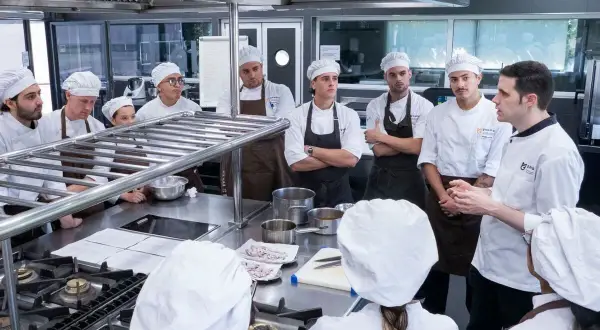 Estudiantes en un curso gestión restaurantes gratis, atentos a las instrucciones de un chef en una cocina profesional.