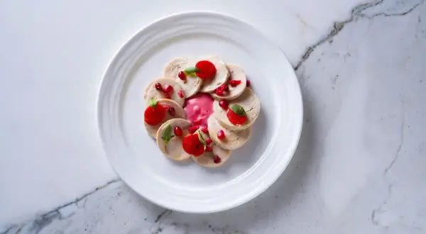 Un delicado plato estrella michelin: finas lonchas de pollo con salsa rosa, granada y hojas verdes, presentado en un plato blanco sobre mármol.