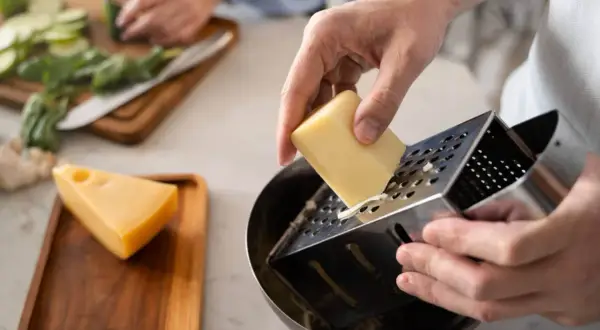 Manos rallando queso para una deliciosa comida. En el fondo, se preparan verduras.